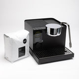 Black coffee machine with a silver spout and a white bag of coffee beans on a light gray background